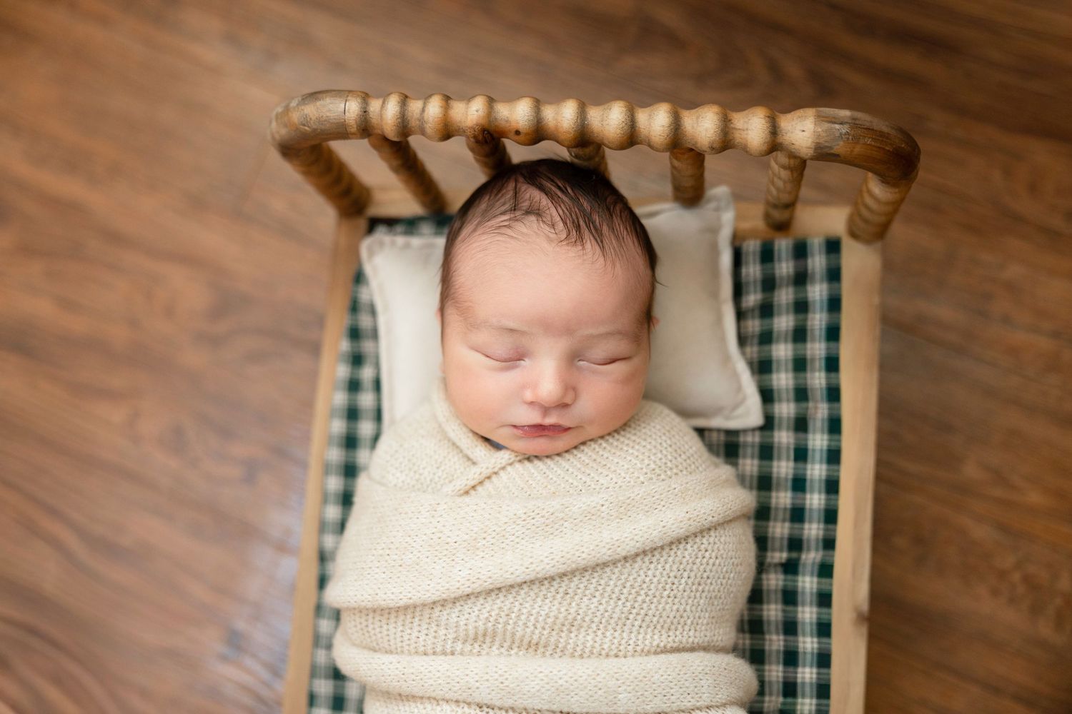 Peaceful newborn sleeping in cream wrap on wooden chair with checkered cushion.