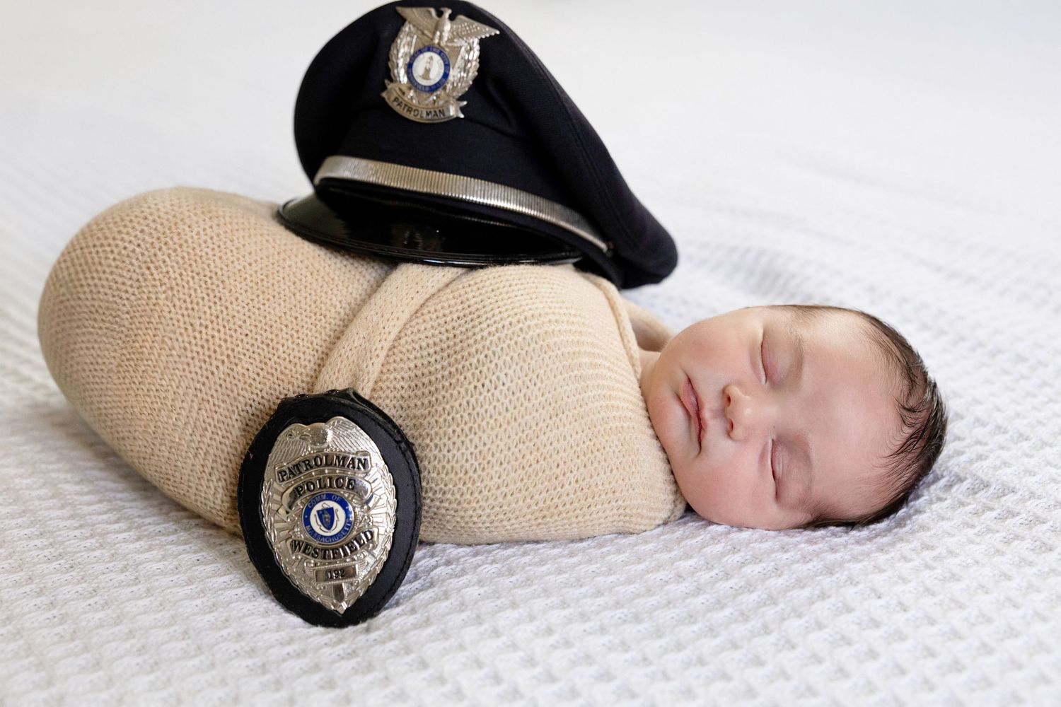 Sleeping newborn wrapped in beige swaddle with police officer hat and badge props on white textured surface.
