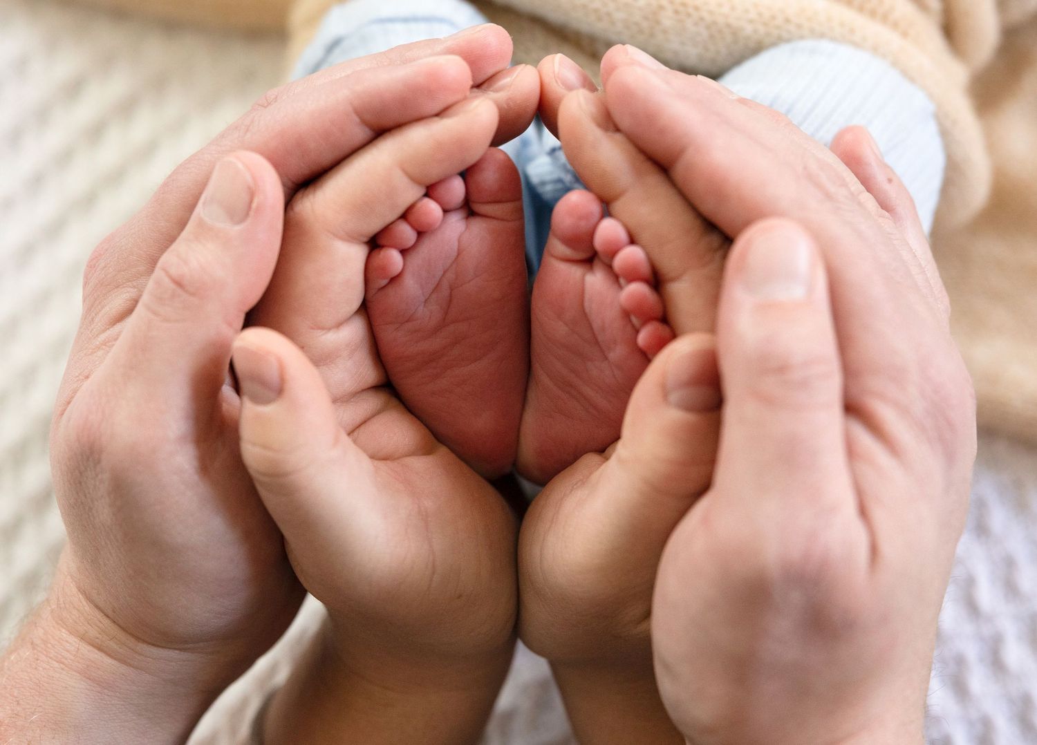 Adult hands gently cradling tiny newborn baby feet against white background.