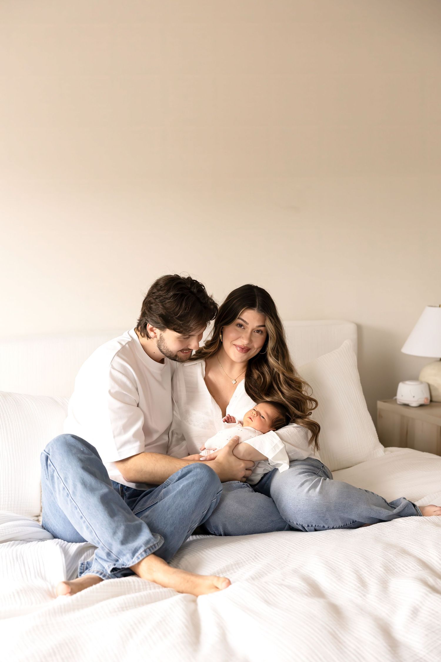 A young couple sitting on a white bed, lovingly gazing at their newborn baby wrapped in a white blanket.