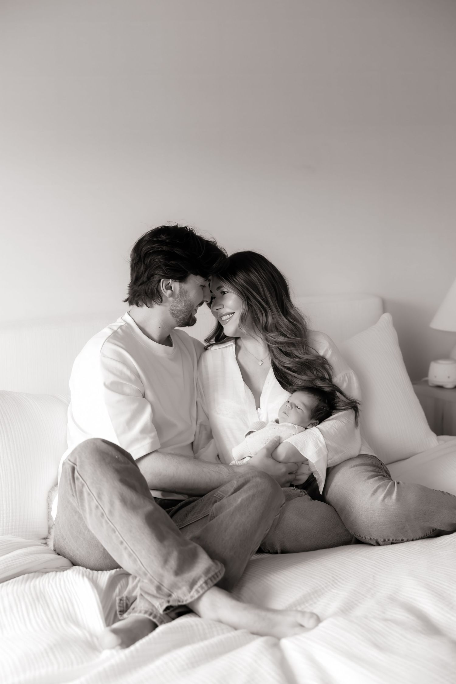 Black and white photo of a couple sitting on a bed, smiling at each other while holding their newborn baby.