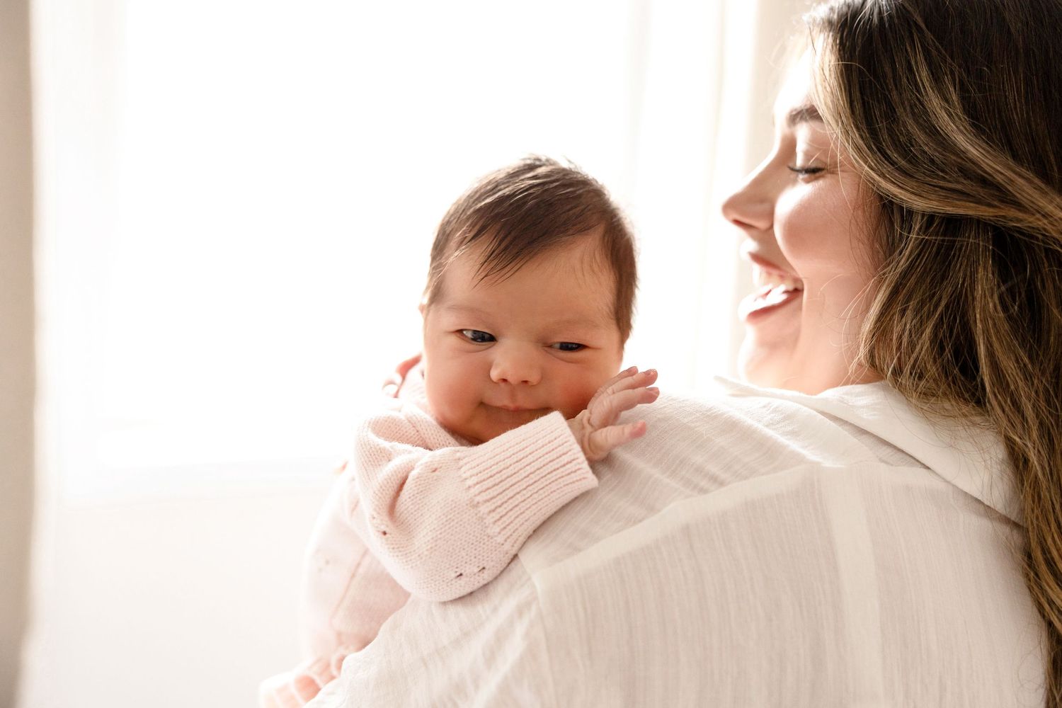 Smiling mother holding her newborn baby dressed in a pink outfit near a bright window.