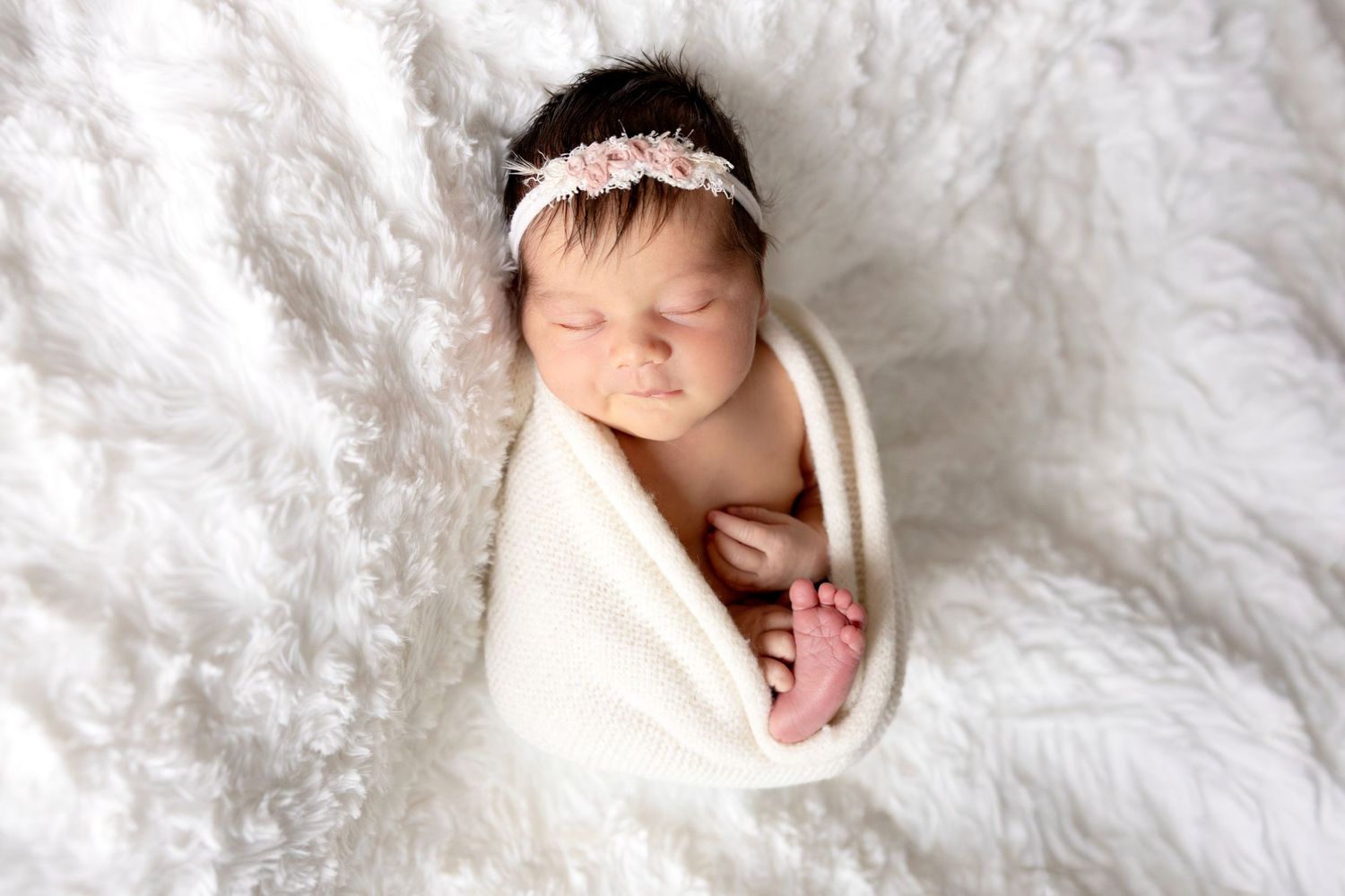 Peaceful newborn baby girl swaddled in white wrap with pink floral headband sleeping on fluffy white blanket.