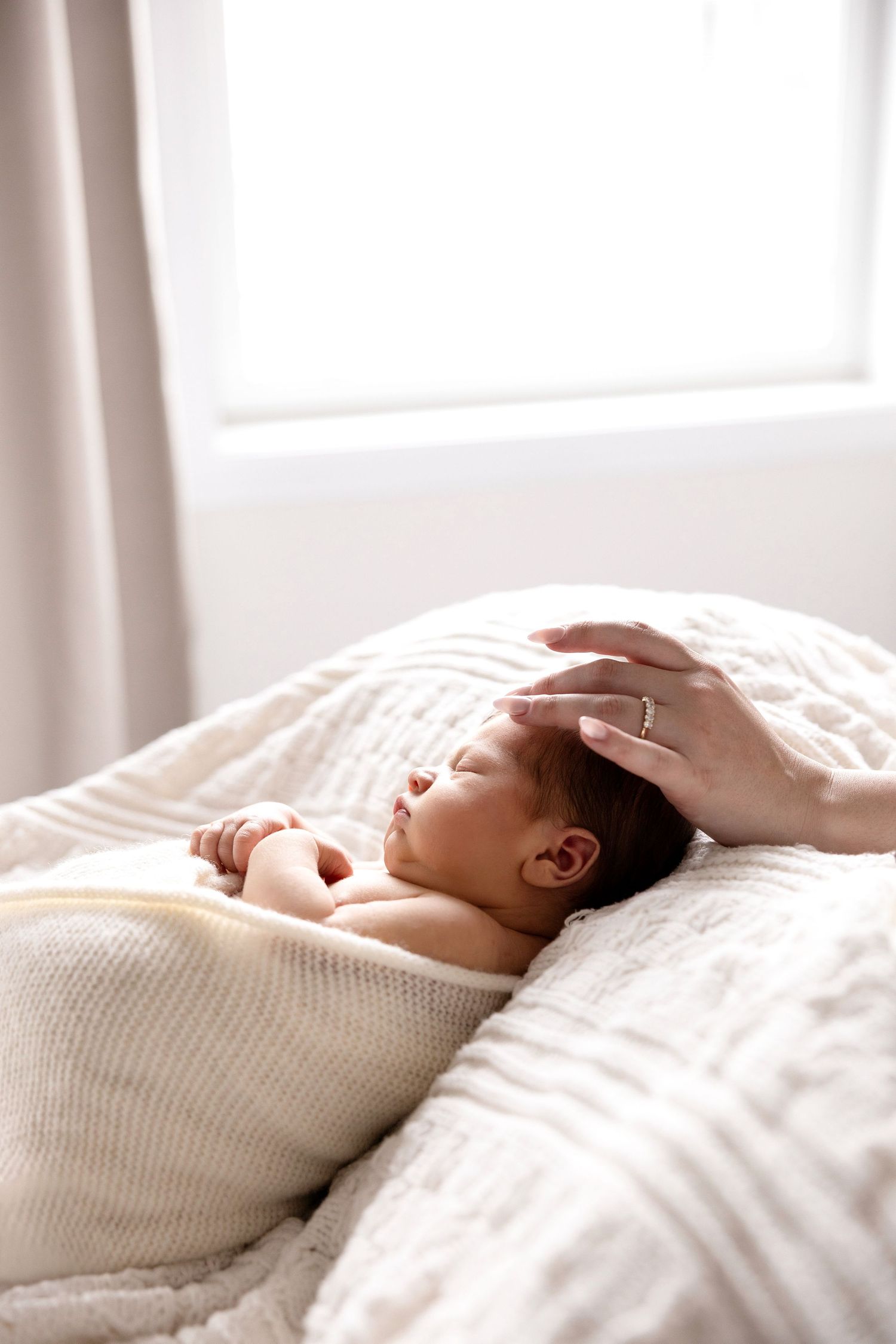 Newborn baby sleeping peacefully on white blanket with mother's hand gently resting on head near bright window.