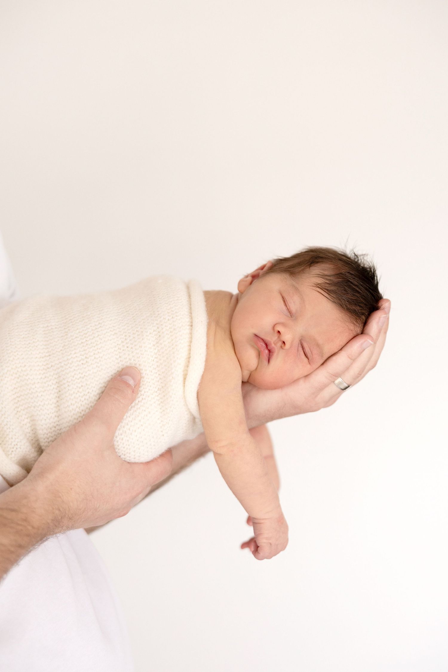 Sleeping newborn baby wrapped in cream knit blanket cradled in father's hands against white background.