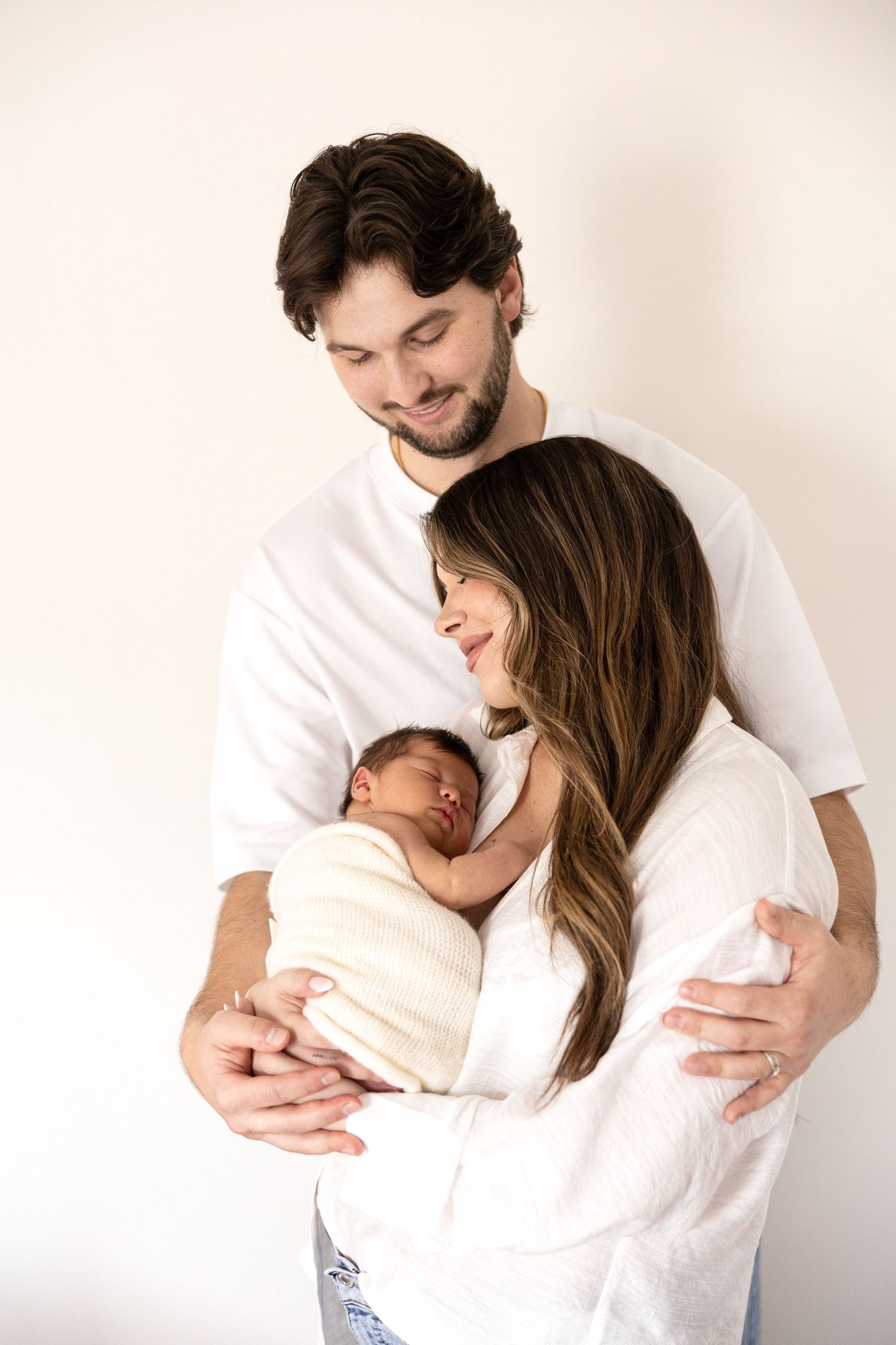 New parents in white clothing lovingly hold their sleeping newborn baby wrapped in a white swaddle.