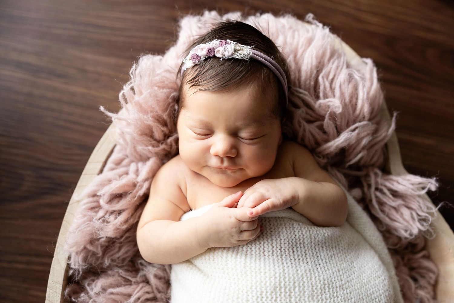 Smiling newborn baby girl in floral headband sleeping peacefully in a round wooden bowl with pink fluffy blanket.