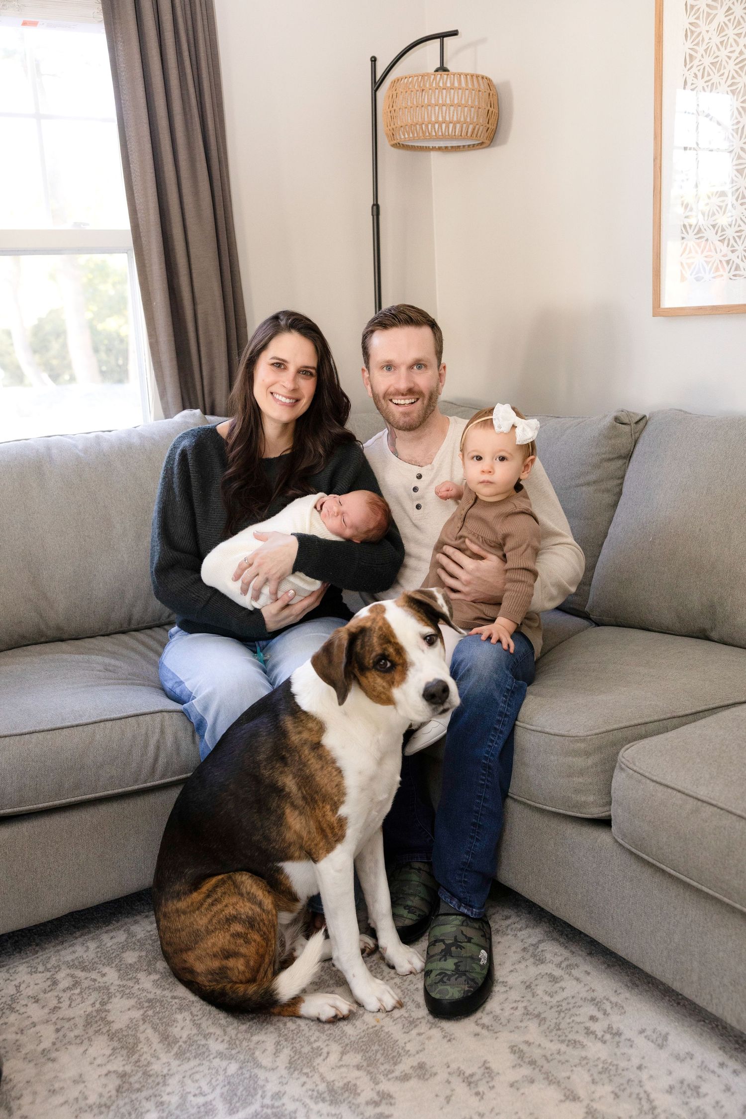 Family portrait on gray couch with their tricolor dog in living room setting.