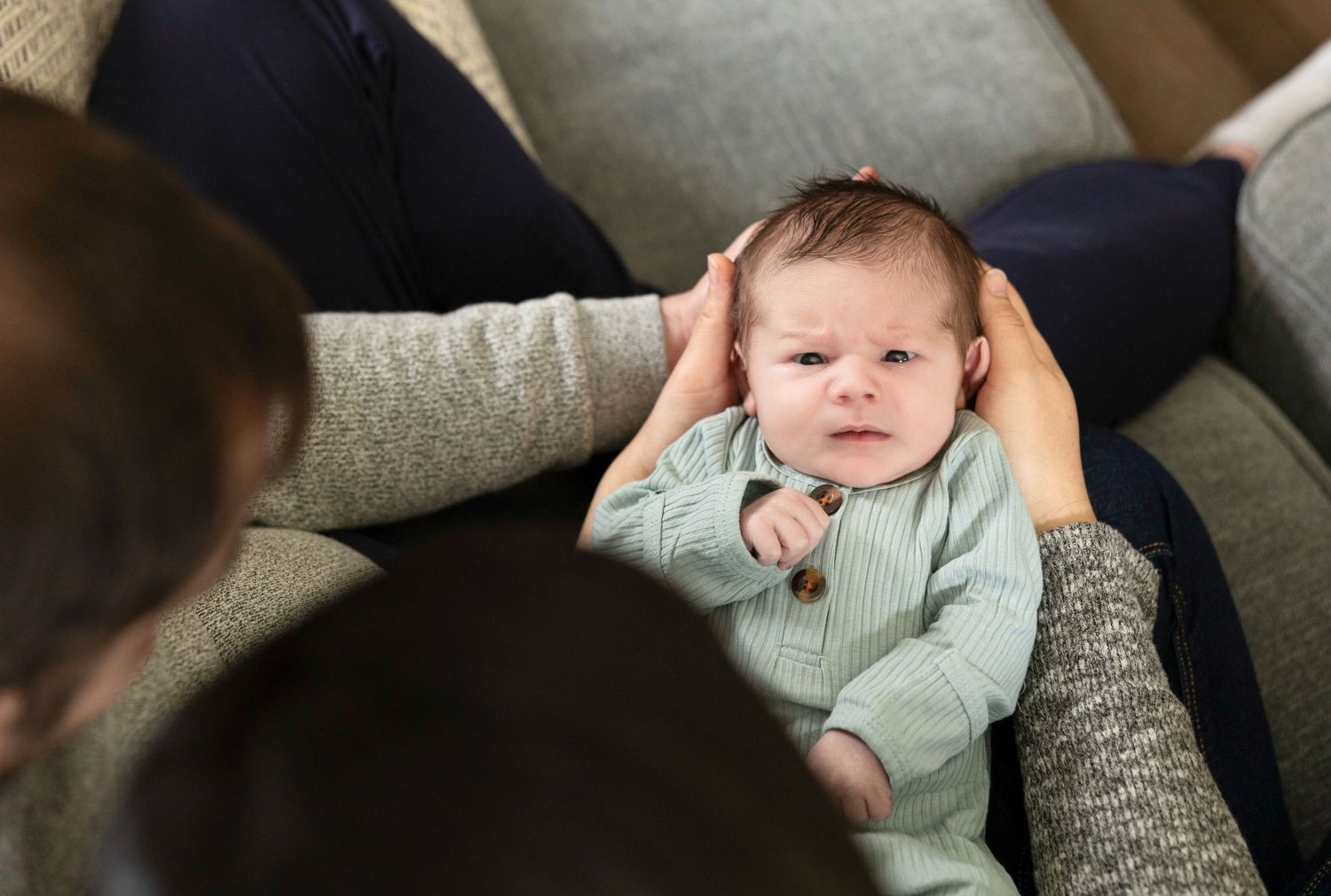 Baby wearing mint green outfit relaxing on grey couch in natural lighting.