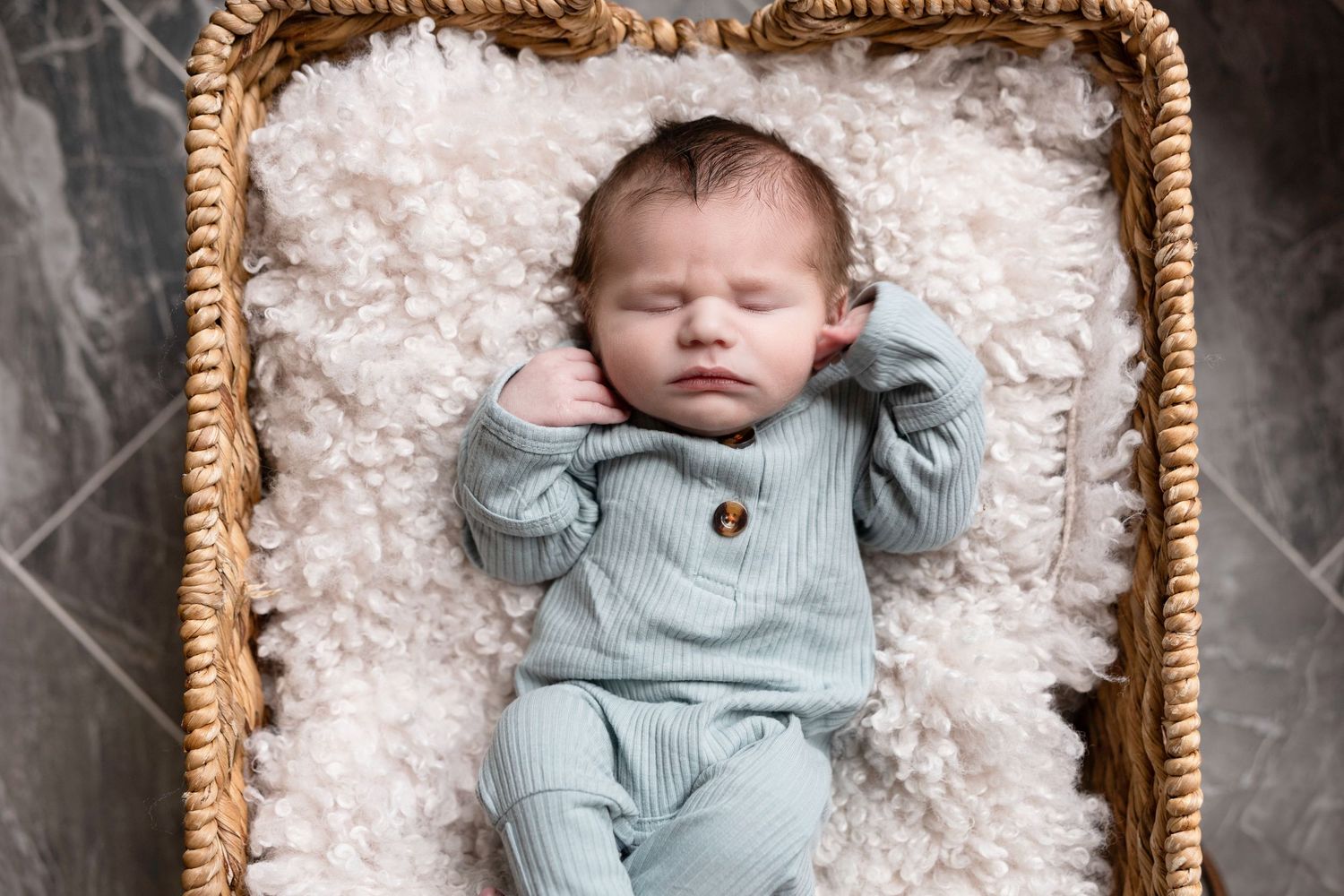 Baby in light blue outfit resting in woven basket on white textured blanket.