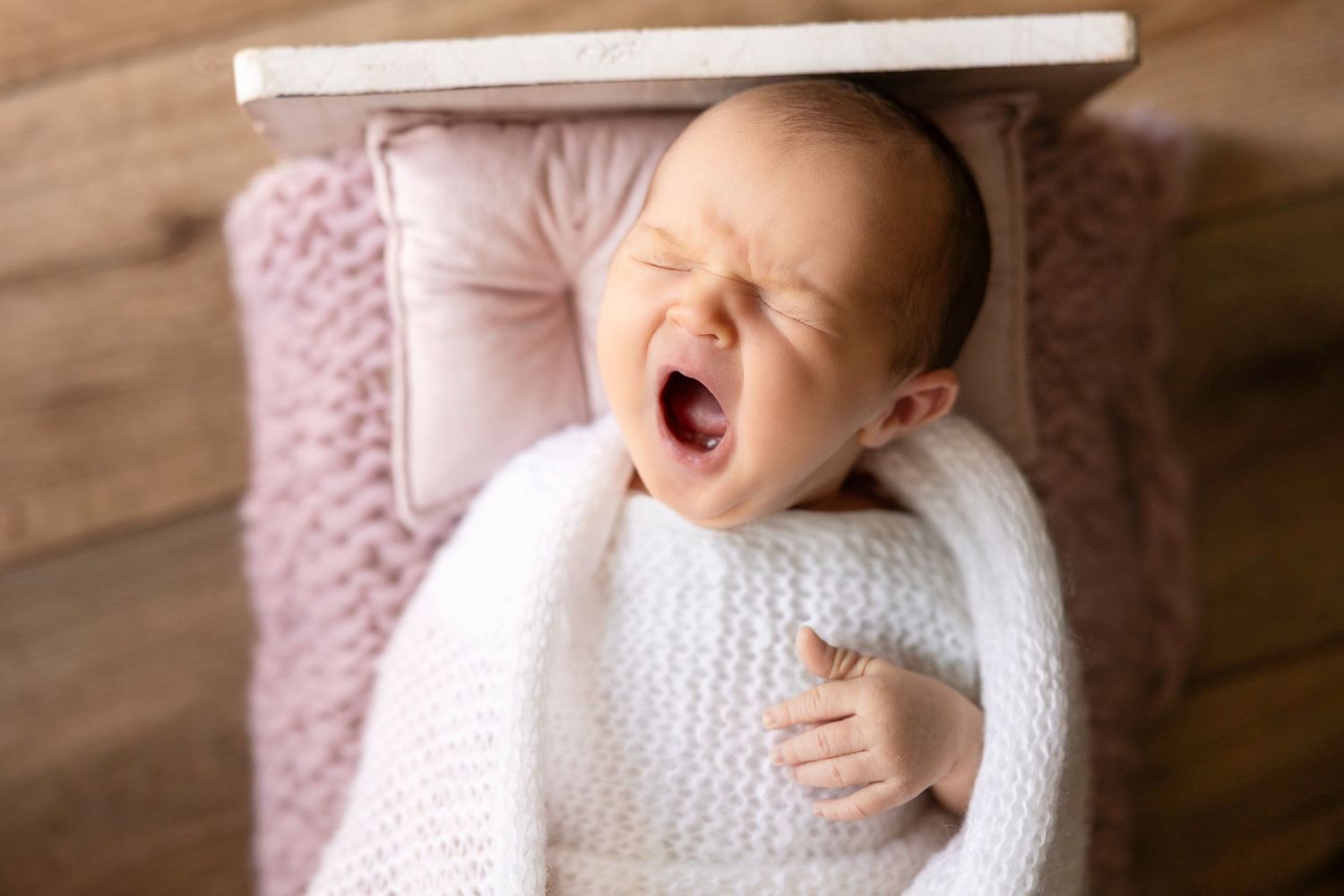 A yawning baby dressed in white lies on a plush pink blanket during a photo shoot.