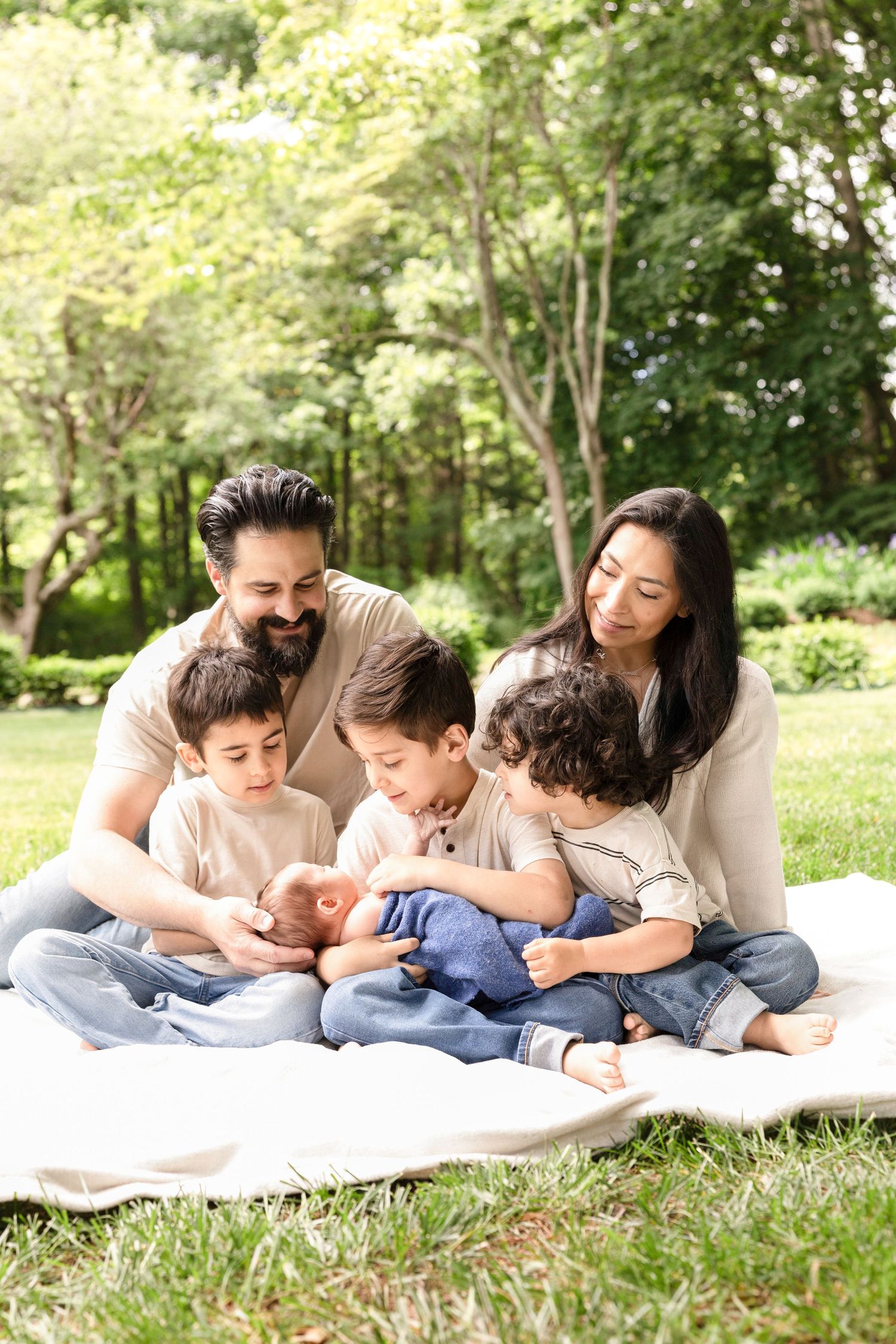 Family relaxing together on white blanket in sunny park setting.