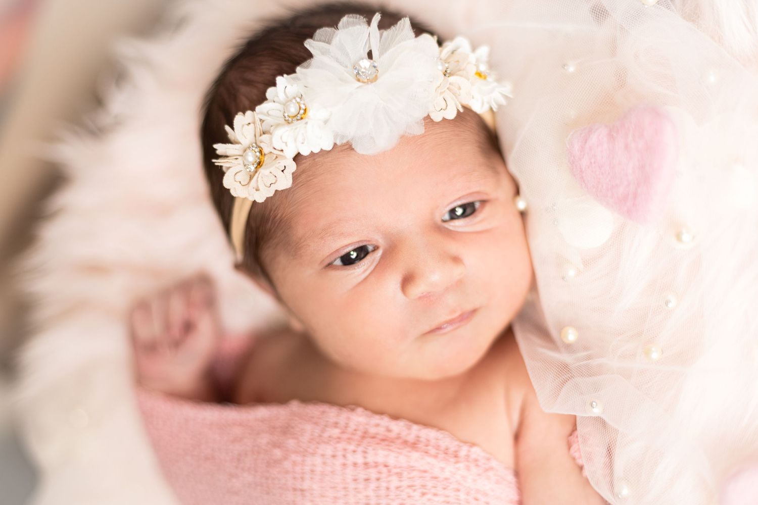 Baby wearing white floral headband against pink background during newborn photoshoot.