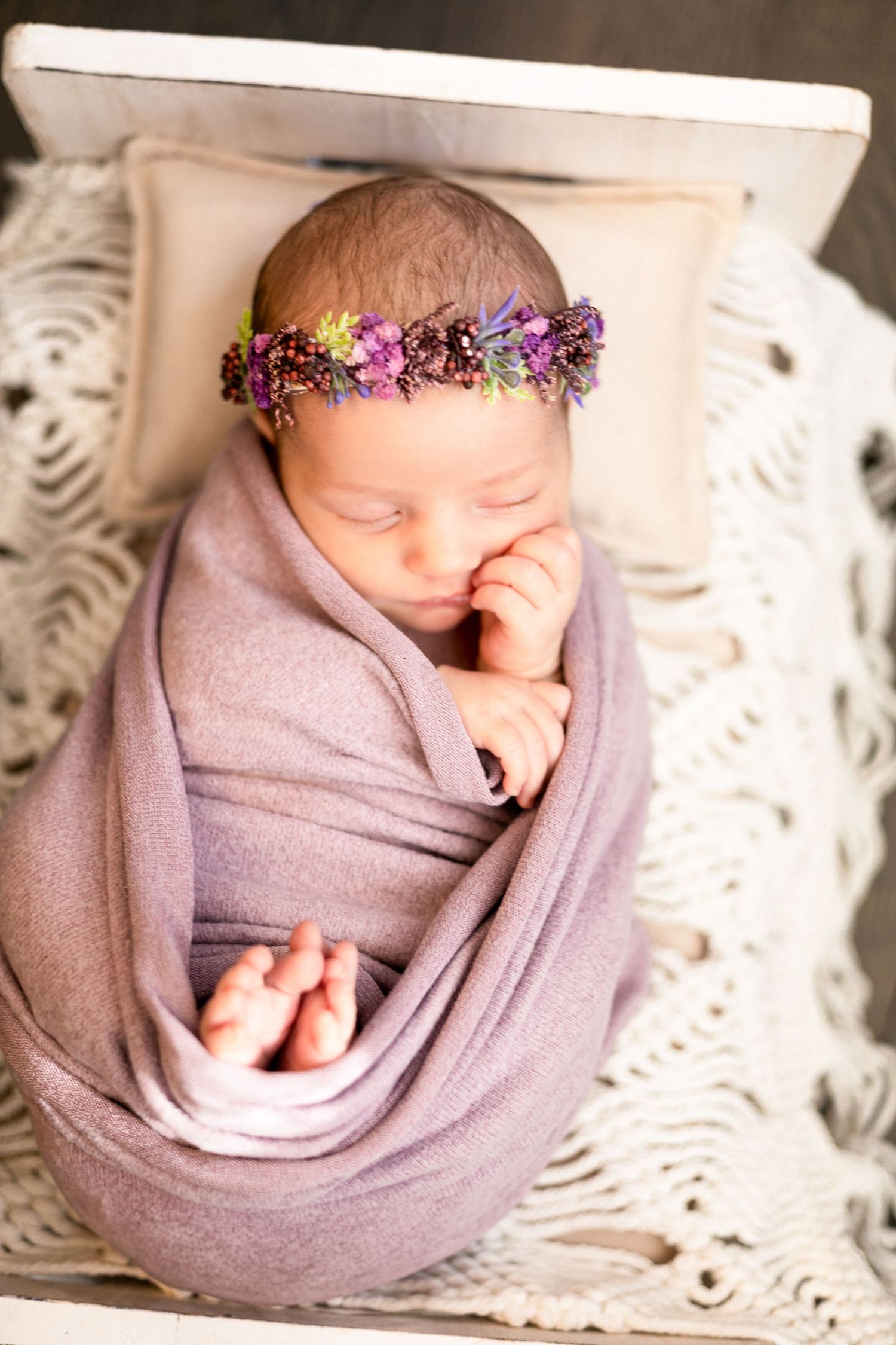 Newborn baby wrapped in purple swaddle with floral crown on textured white backdrop.