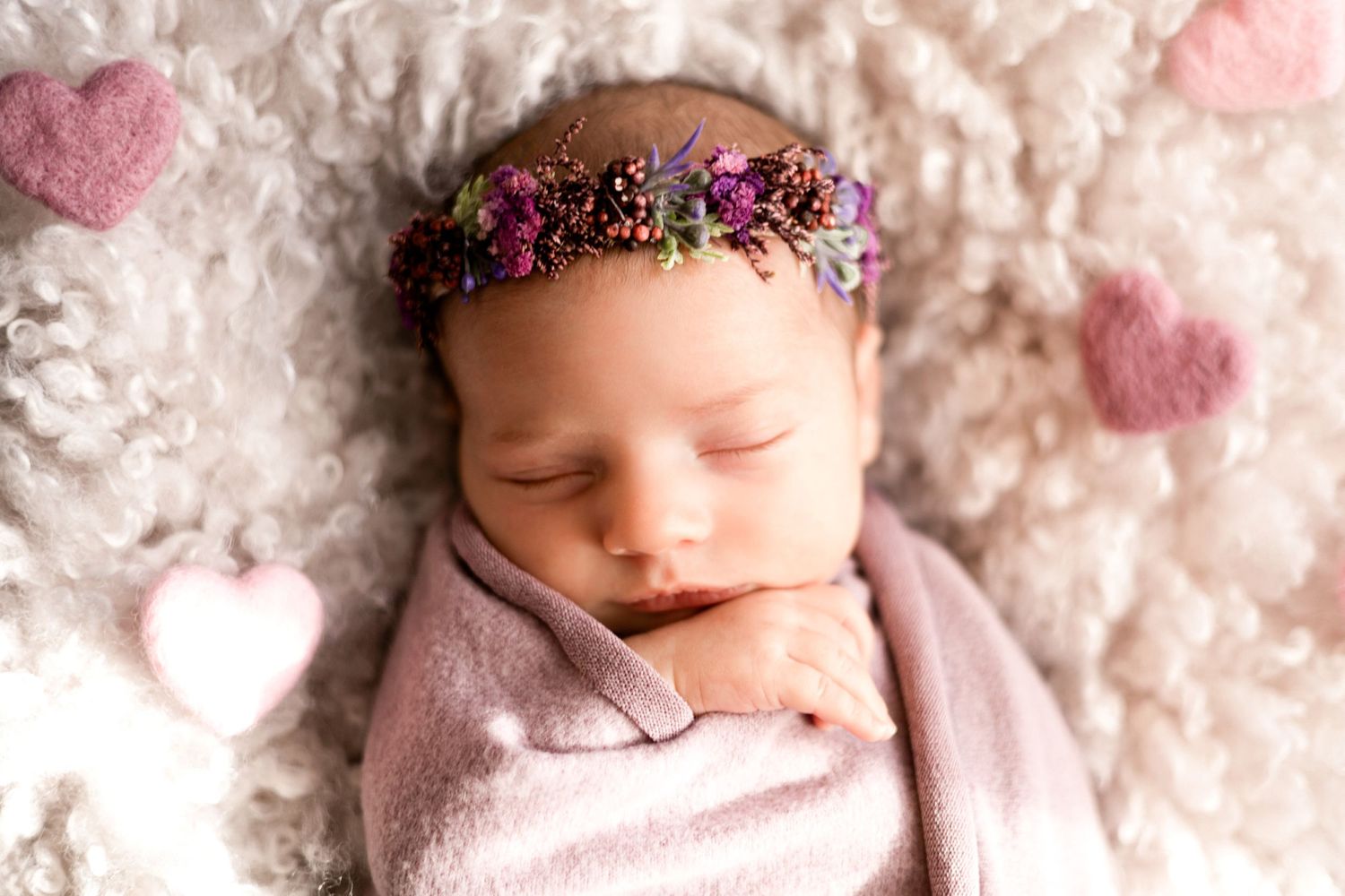 Sleeping newborn in pink wrap with delicate floral crown against white texture.