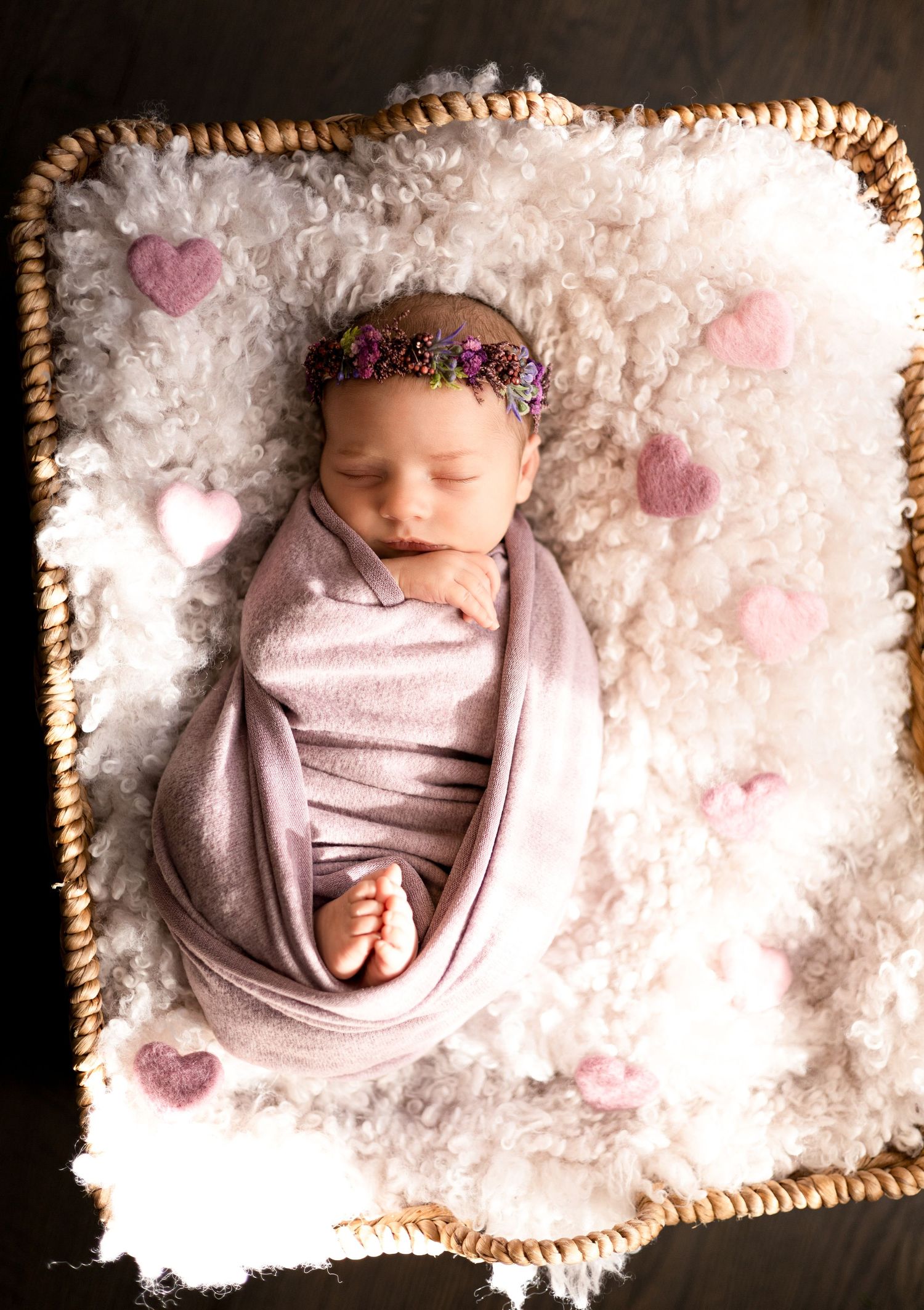 Newborn baby in basket surrounded by white fluffy texture and pink heart decorations.
