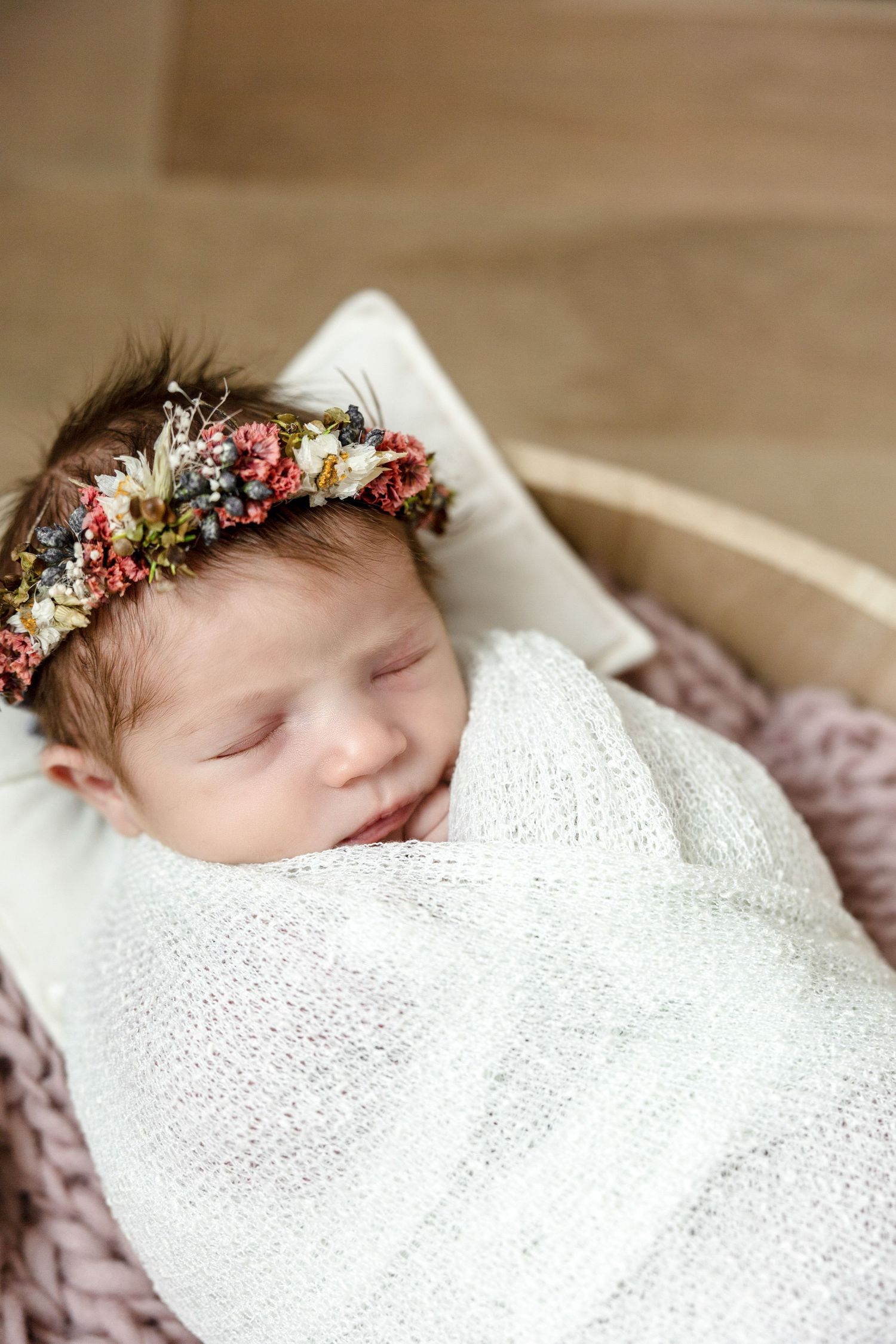 Sleeping newborn wrapped in white blanket wearing delicate floral crown.