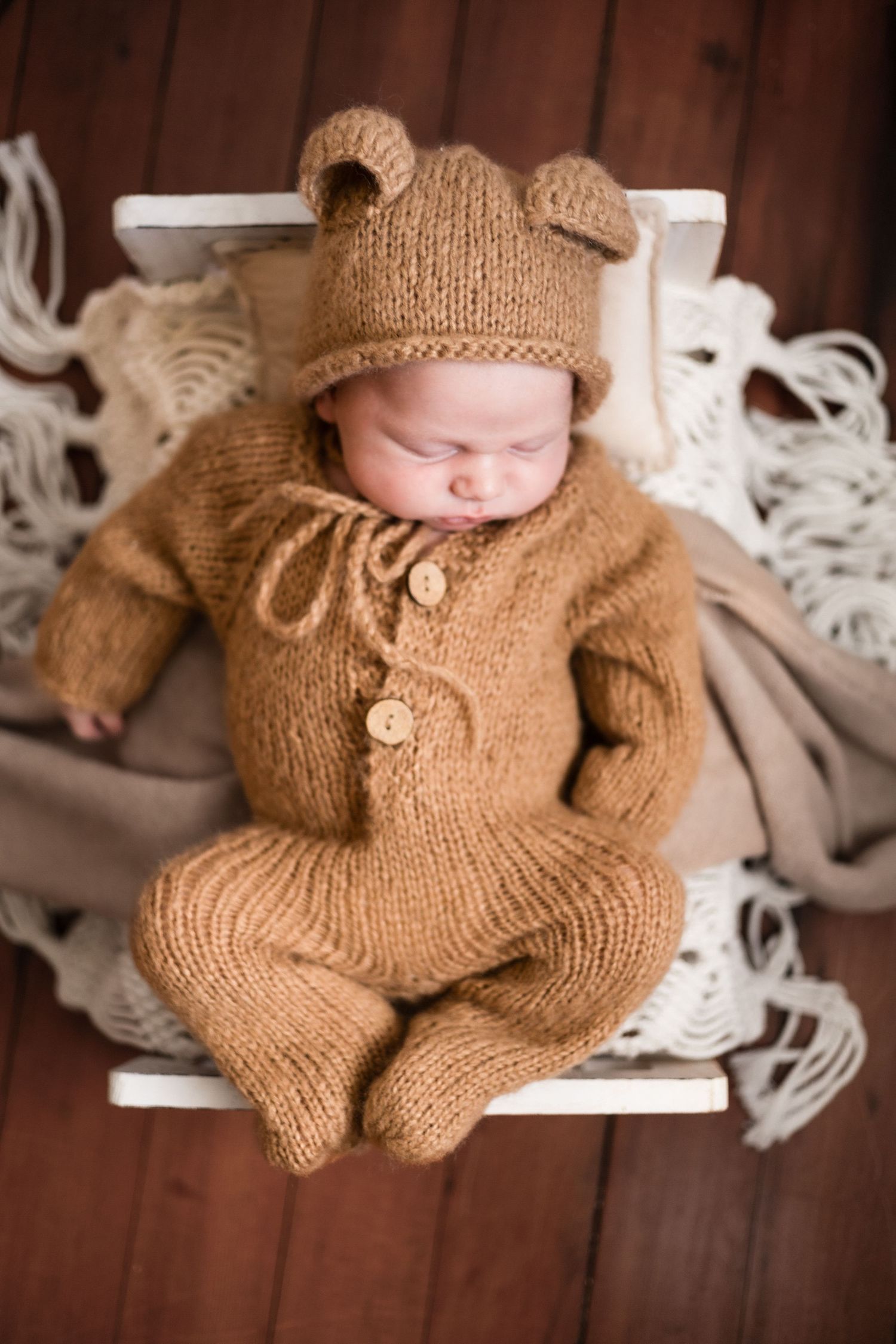 Newborn wearing knitted tan outfit photographed on white lace backdrop.