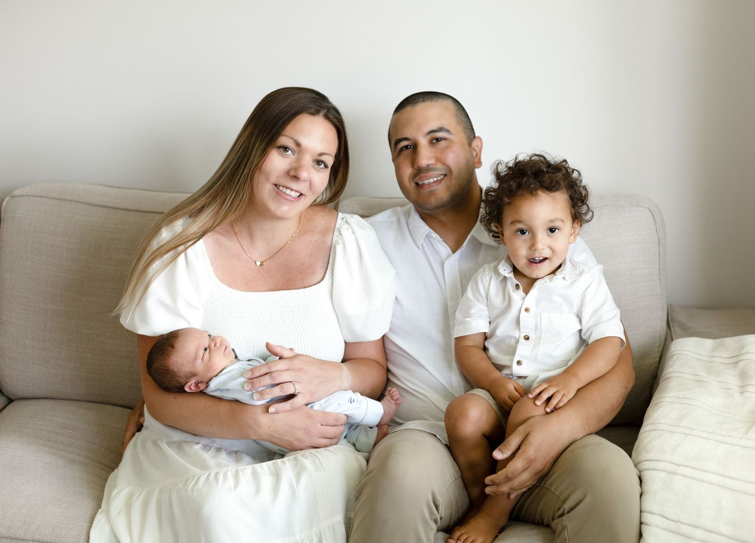 Family lifestyle session on white couch with newborn and toddler in casual attire.