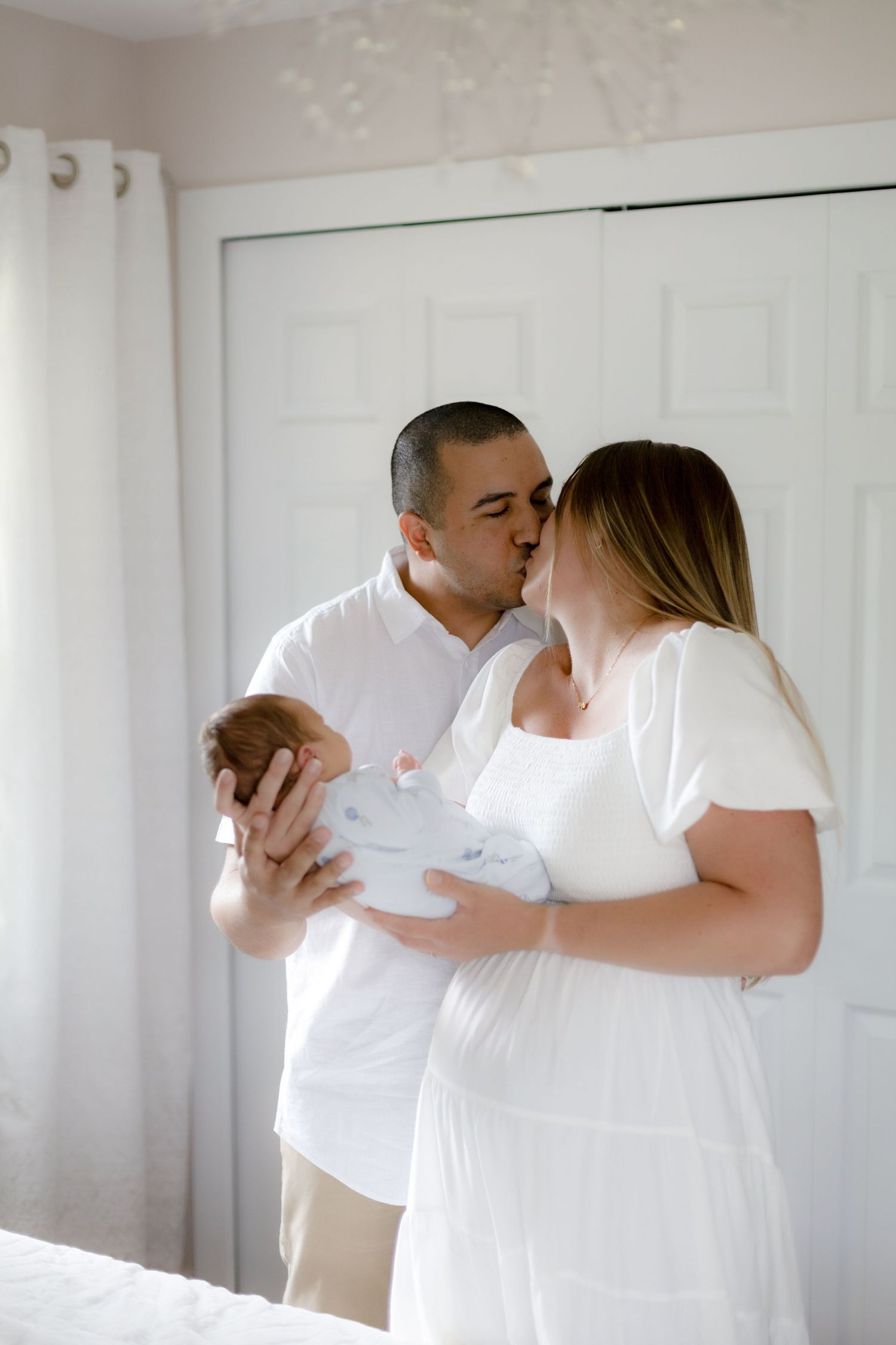 A tender family moment as parents in white outfits share an intimate kiss while holding their newborn baby.