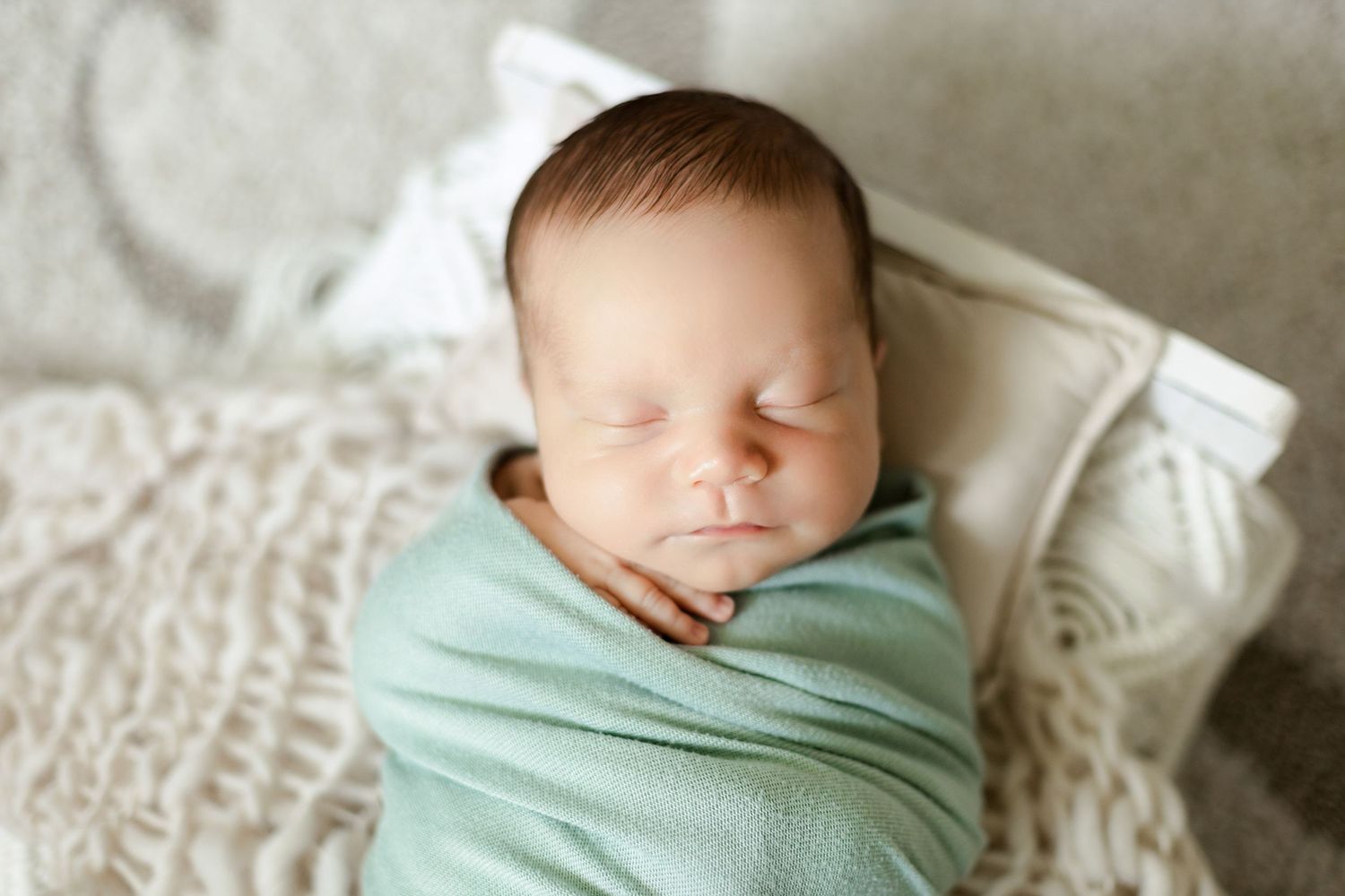 Sleeping newborn baby wrapped in mint green swaddle on textured cream blanket.