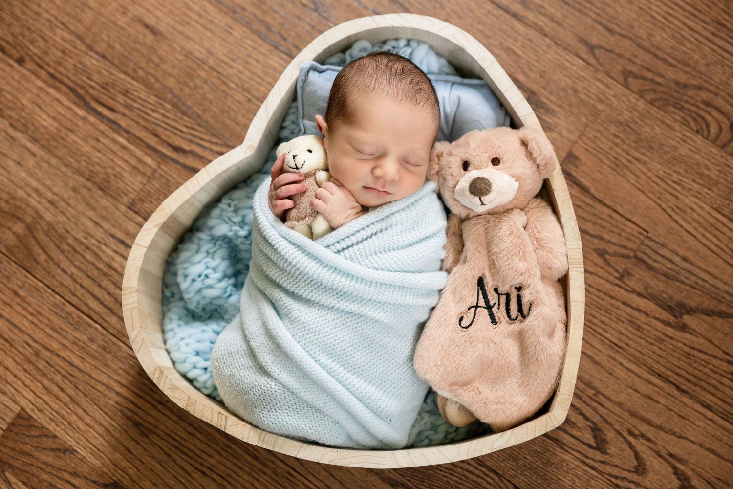 Newborn baby sleeping in heart-shaped basket with blue wrap and teddy bear.