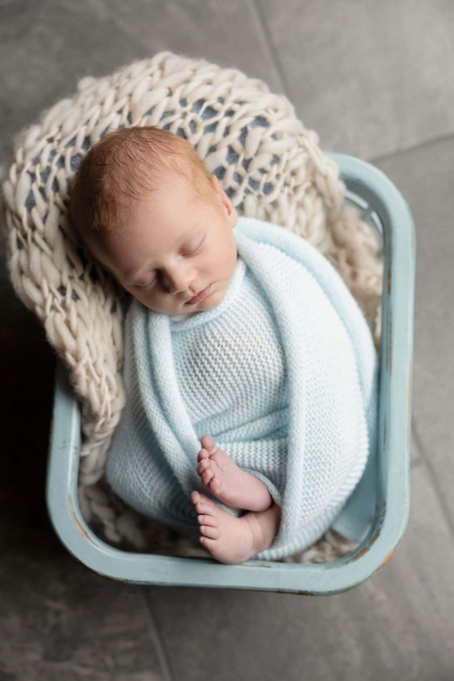 Newborn baby sleeping in light blue knit wrap in vintage blue basket.