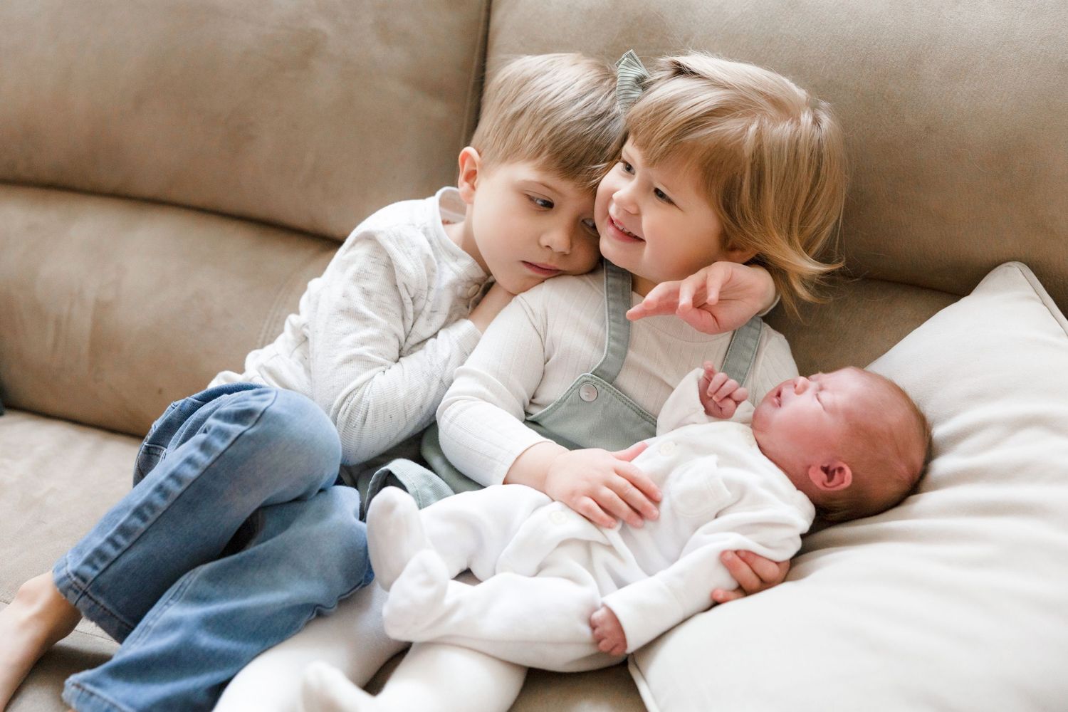 Young siblings cuddle together on a beige couch with their newborn sibling.