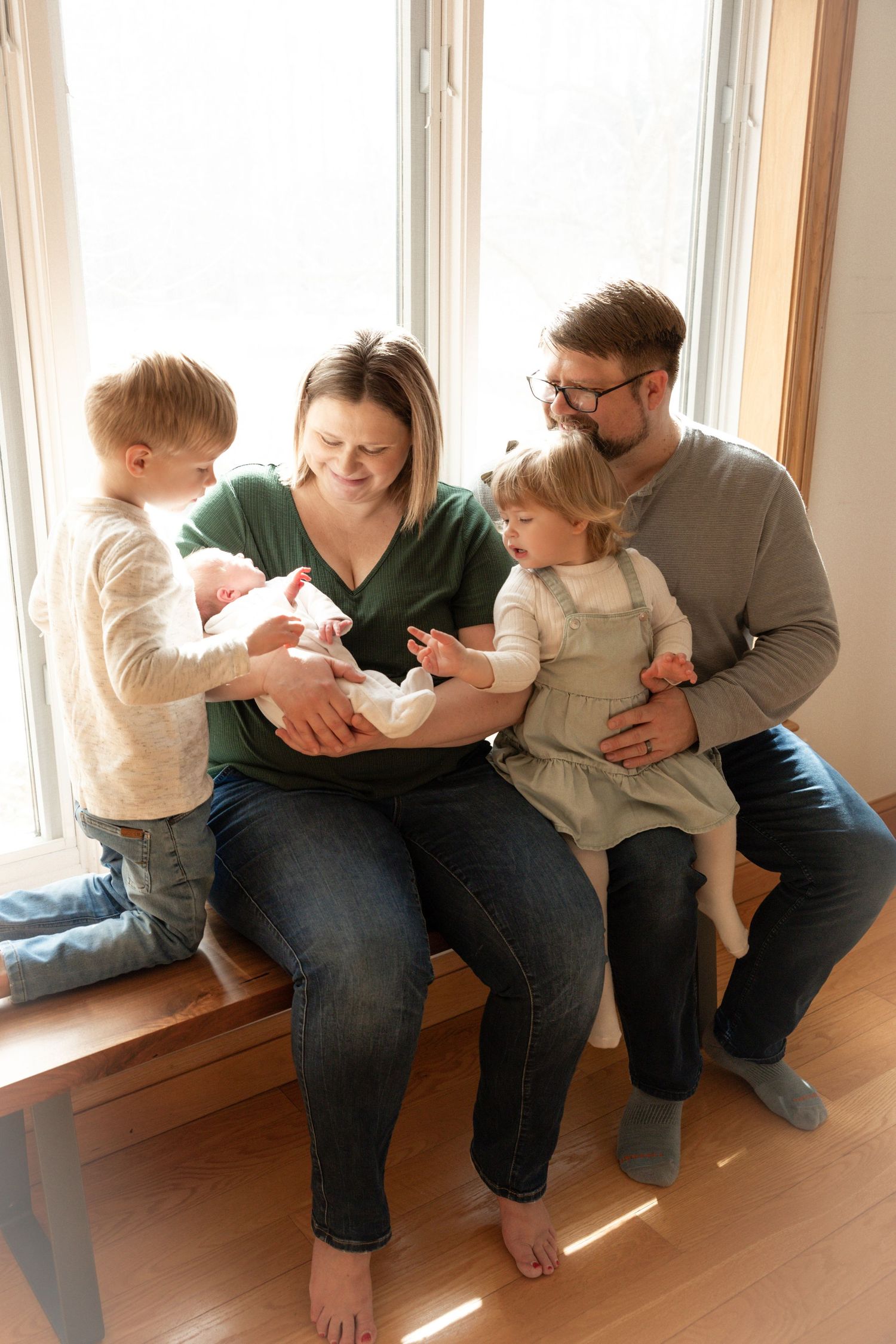 Family sits together on window bench sharing intimate moments with new baby.