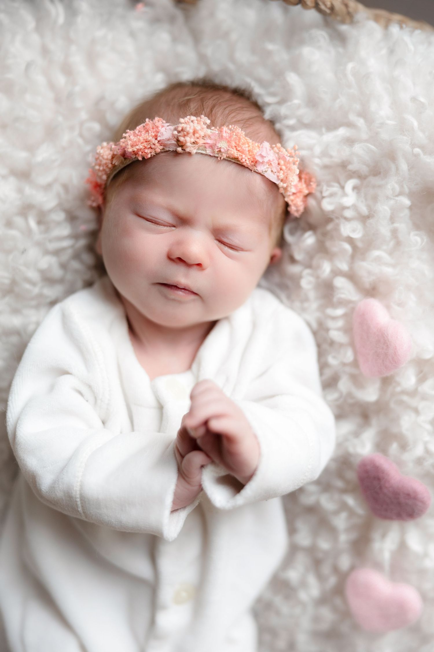 Sleeping newborn in white outfit wears peach headband on fluffy white blanket.