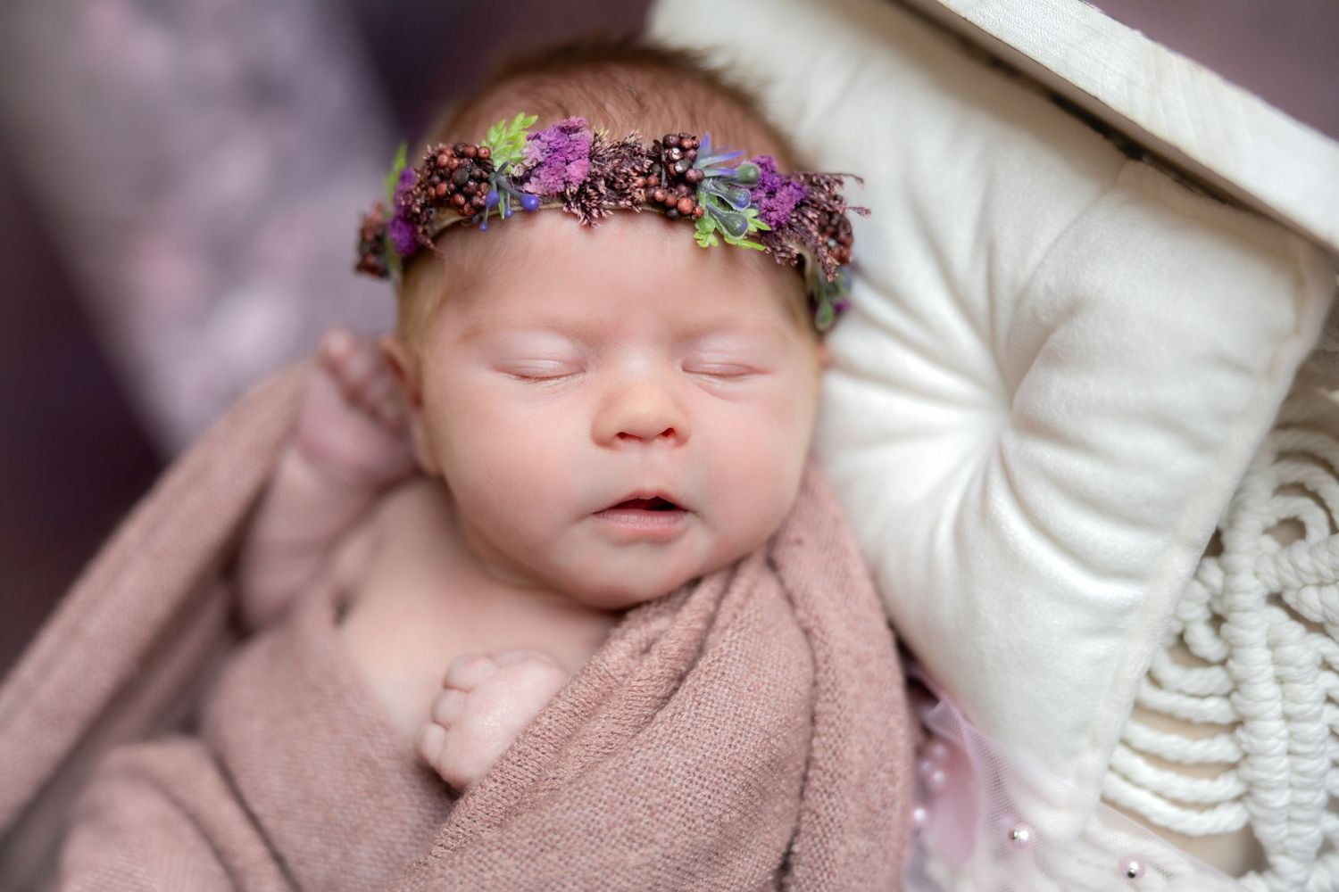 Peaceful newborn sleeps wearing purple floral headband and wrapped in pink blanket.