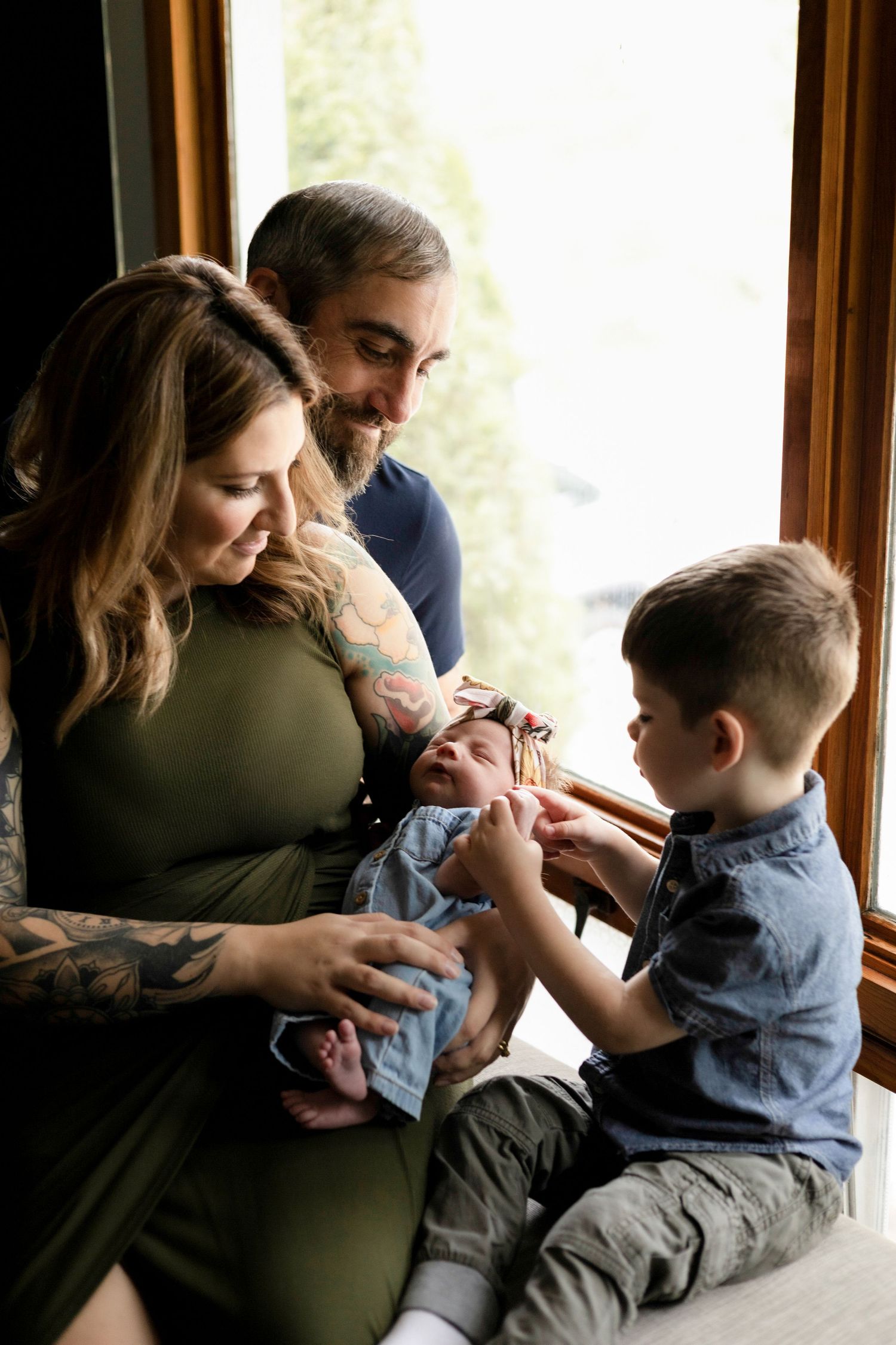 Family bonding moment by window as they cuddle with their newborn and toddler.