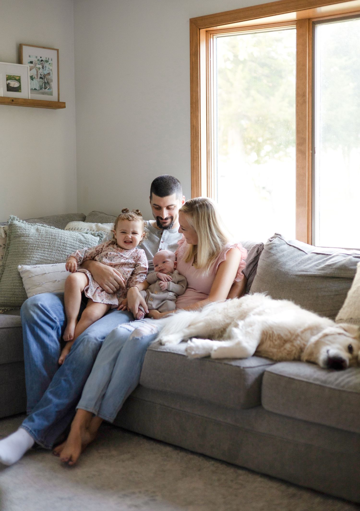 Family relaxes together on grey sectional sofa with white dog in sunny living room.