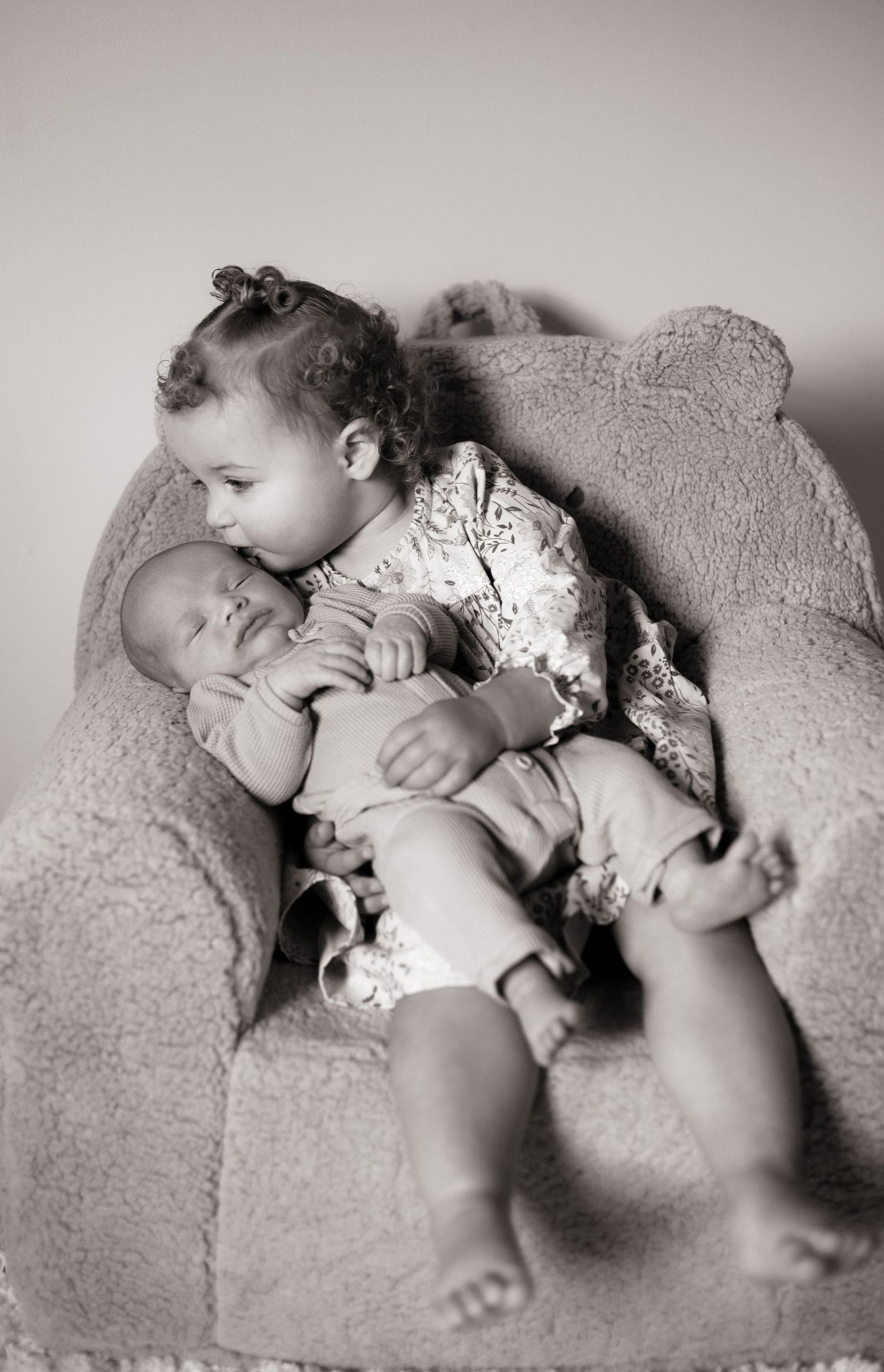 Two young children napping together on plush grey couch in black and white photo.