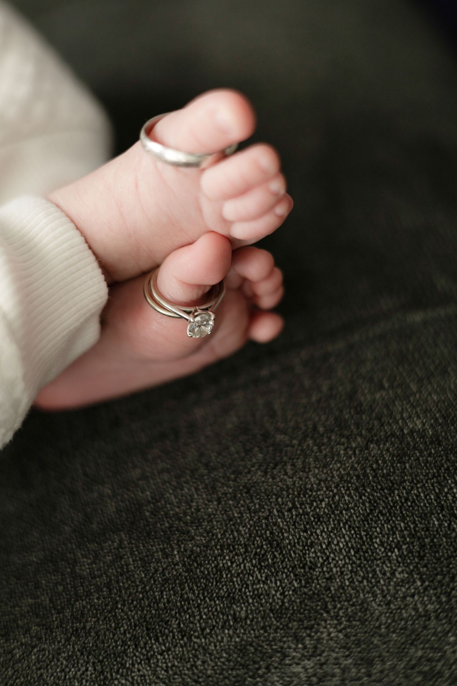 Close-up shots of tiny baby fingers grasping a delicate diamond engagement ring on dark fabric.
