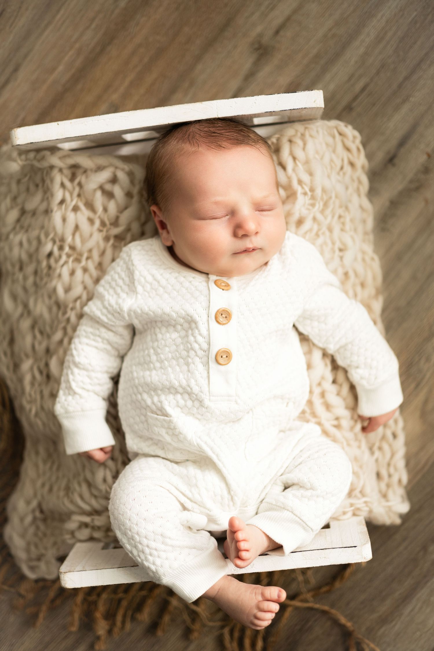 Newborn baby dressed in white knit outfit lying on textured cream blanket with wooden backdrop.