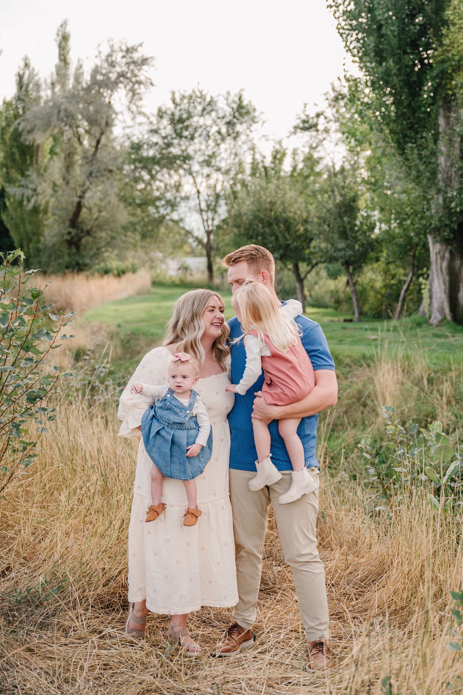 A couple stand together in a field of tall grass at sunset, each holding a small child in their arms.