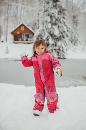 Small child in pink snowsuit plays in deep winter snow near cabin.