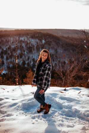 Person in plaid jacket poses on snowy mountain overlook at sunset.