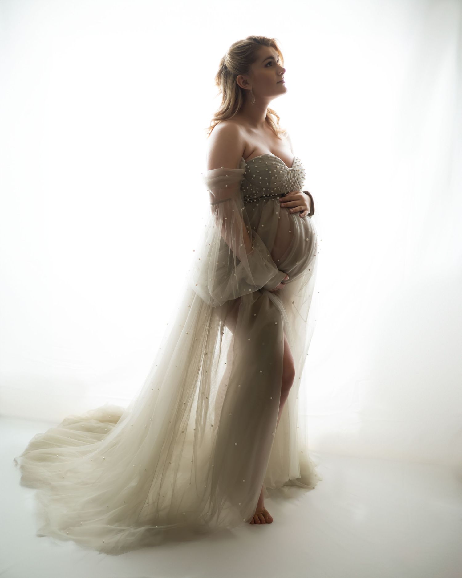Ethereal maternity portrait in flowing white dress against bright background.