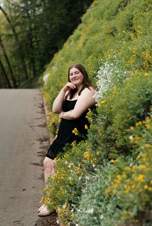 A person in a black dress poses along a winding path bordered by vibrant yellow and white wildflowers on a sunny day.