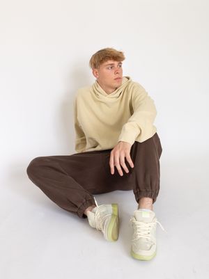 Person in beige sweater and brown pants sitting casually on floor wearing cream and yellow sneakers against white background.