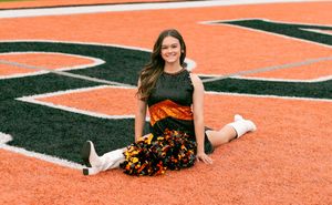 Cheerleader in black and orange uniform sits on orange and black turf field during photo session.