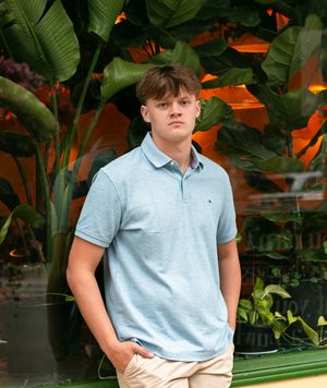 Someone in a light blue polo shirt and khaki shorts poses against a backdrop of tropical plants with orange lighting.