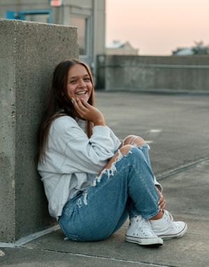 A casual streetwear look with distressed jeans, white sweater and converse shoes against a concrete wall at sunset.
