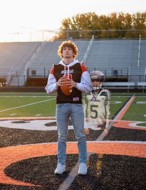 High school athlete in a varsity jacket and jeans poses with a football on a school field at sunset.