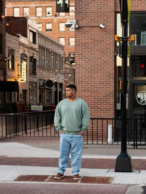 Person in mint green sweater and light jeans standing on brick sidewalk in an urban setting with historic buildings.