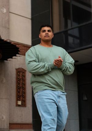 A person wearing a mint green sweatshirt and light blue jeans stands and poses against a building exterior.