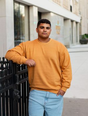 Person in orange sweatshirt and light denim jeans leans against black metal fence outside building.