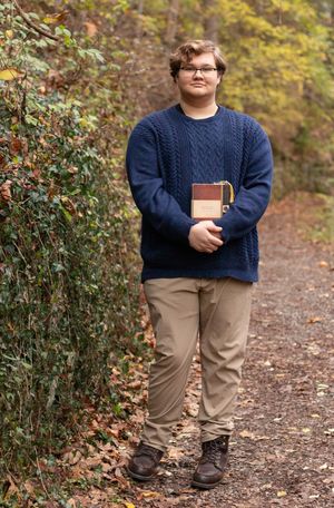 Student in navy cable knit sweater holds book while standing on autumn forest trail with fallen leaves.